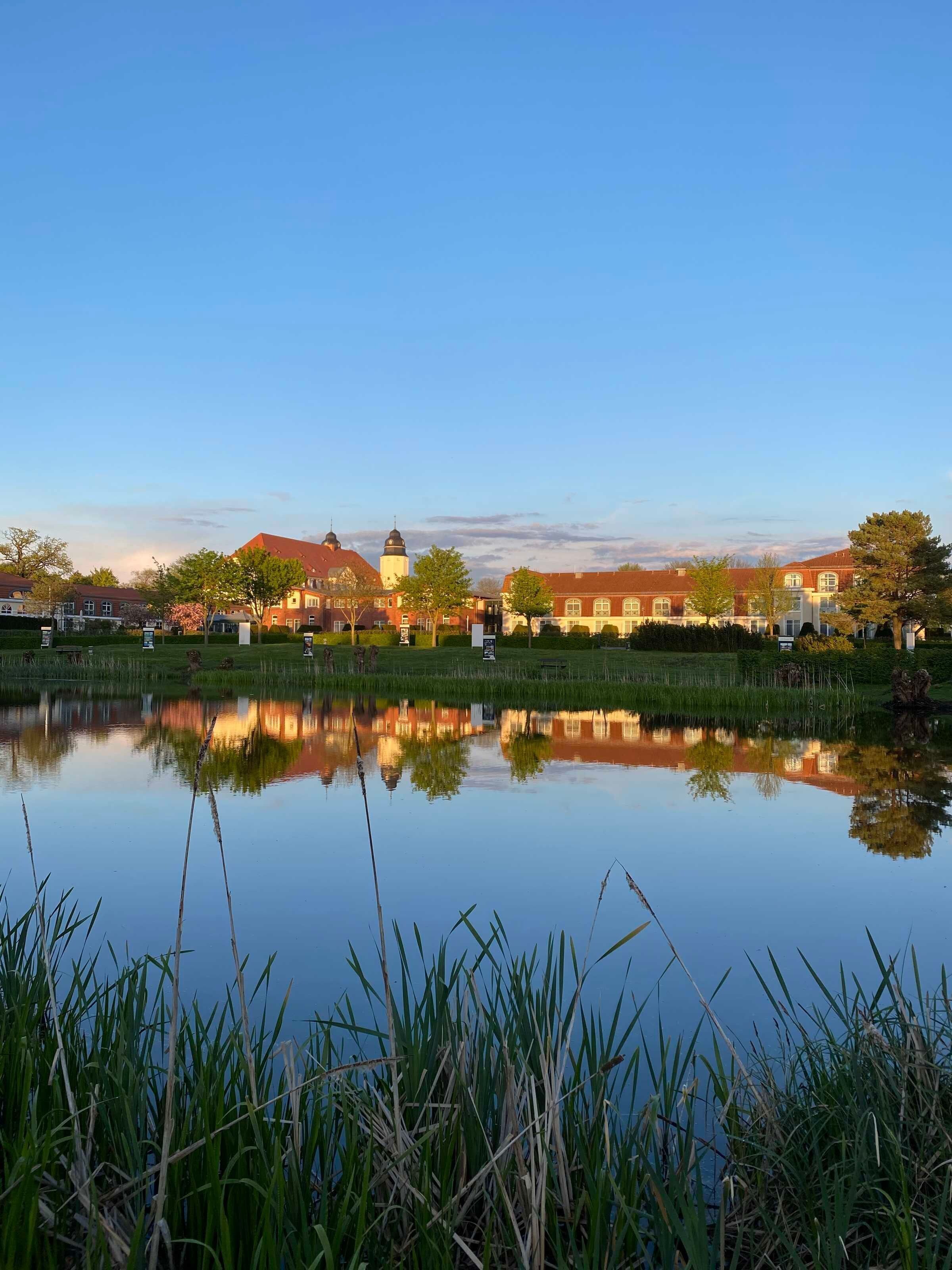 A serene lakeside view with a reflection of buildings and trees, a clear sky, and a calm water surface.