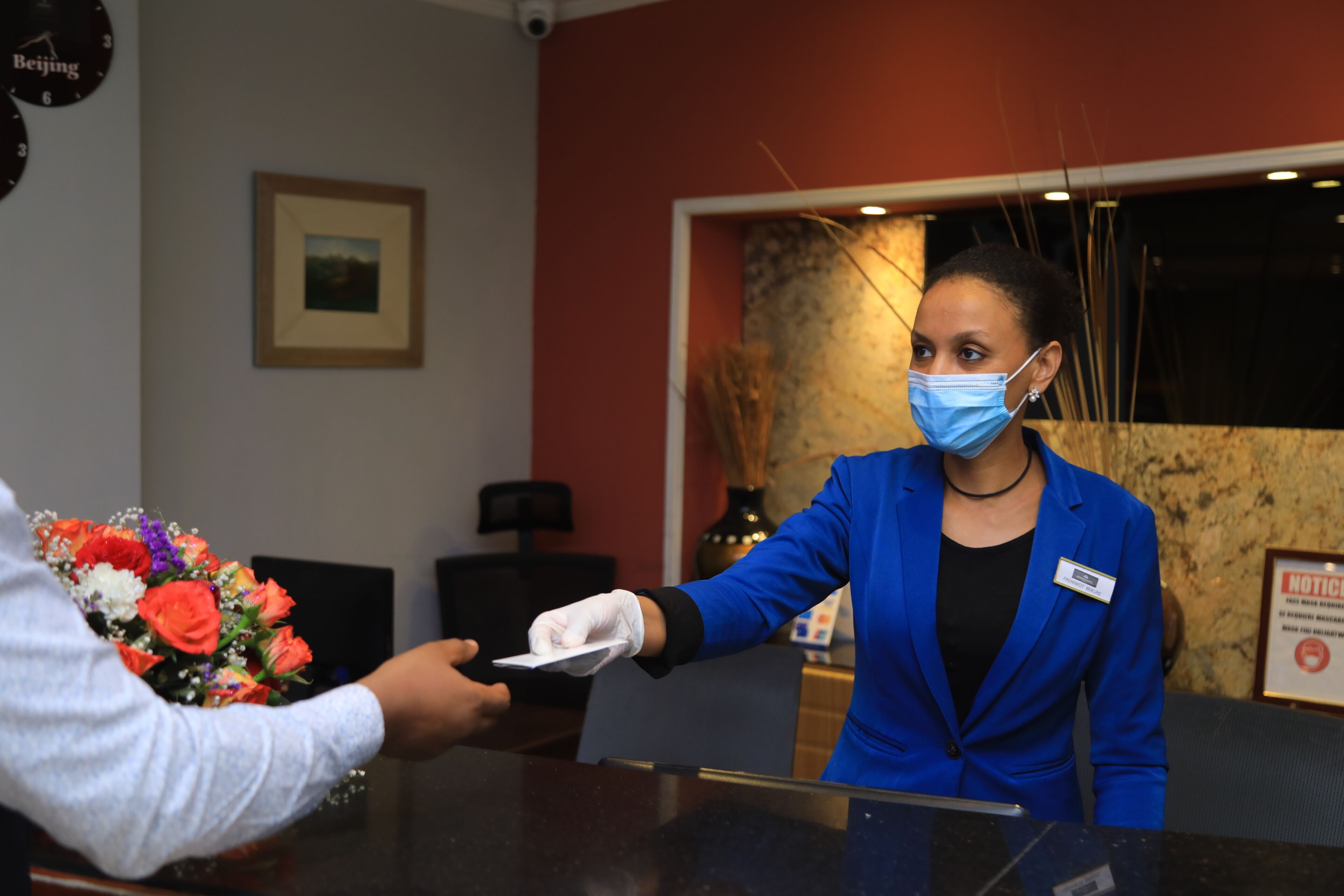 Een receptiebalie met een receptionist in een blauw colbert. Er hangt een lijst op de muur en een klok met de tekst 'Beijing'.