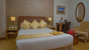Habitación de hotel con una cama grande, dos mesitas de noche con lámparas, una mesita auxiliar de madera, una silla, una pequeña mesa con un florero y un cuadro en la pared.
