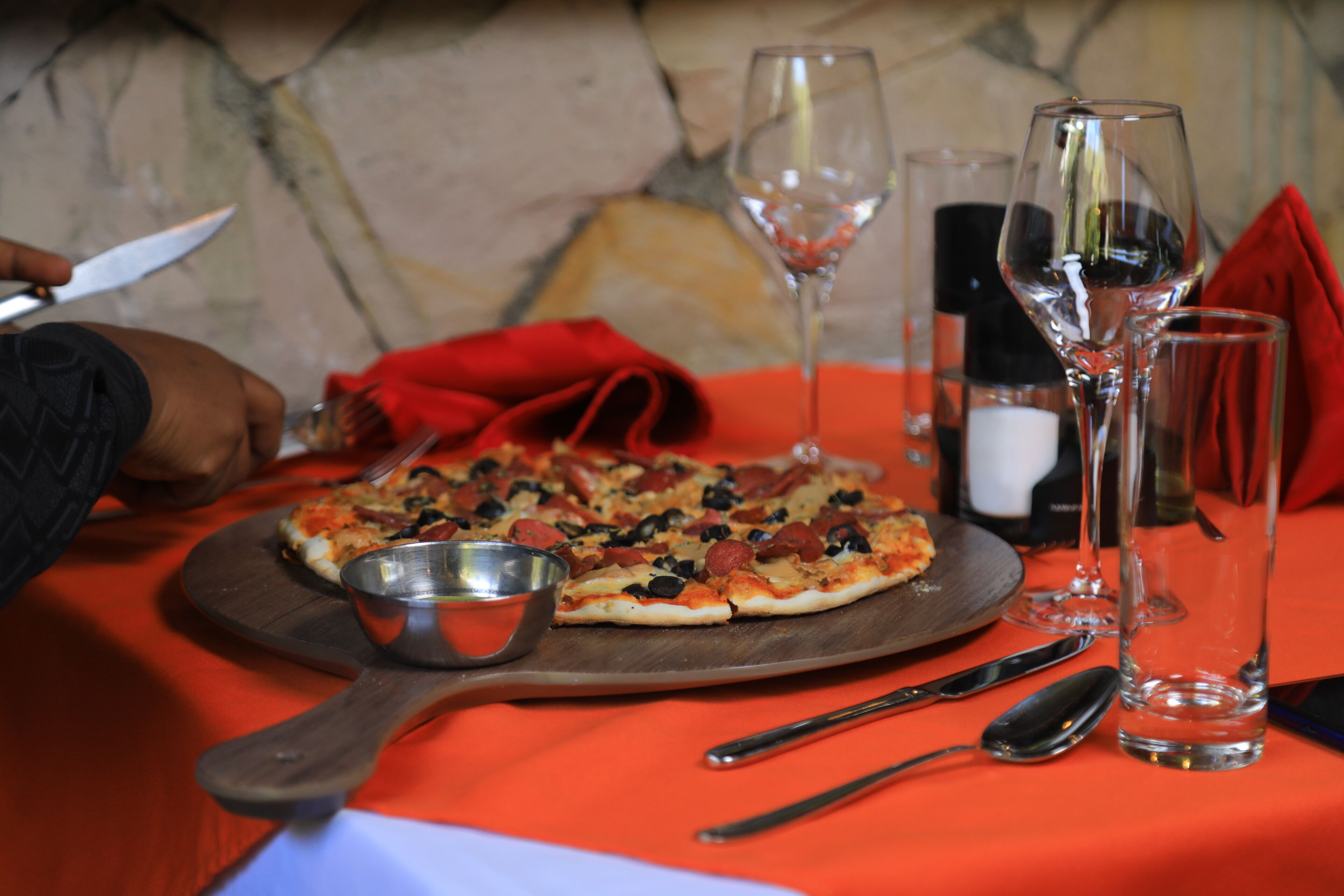 A pizza on a wooden board with a metal serving dish, a knife, and a spoon on a table with a red tablecloth.