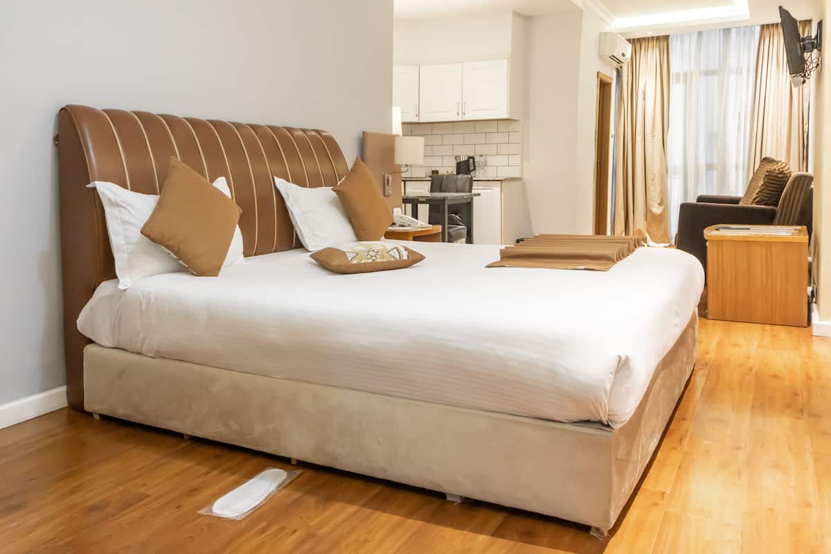 A hotel room with a large bed, a wooden floor, a desk with a chair, a kitchenette, and a TV mounted on the wall.