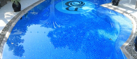 A large, clear blue pool with a tiled pattern, surrounded by potted plants and outdoor seating areas.