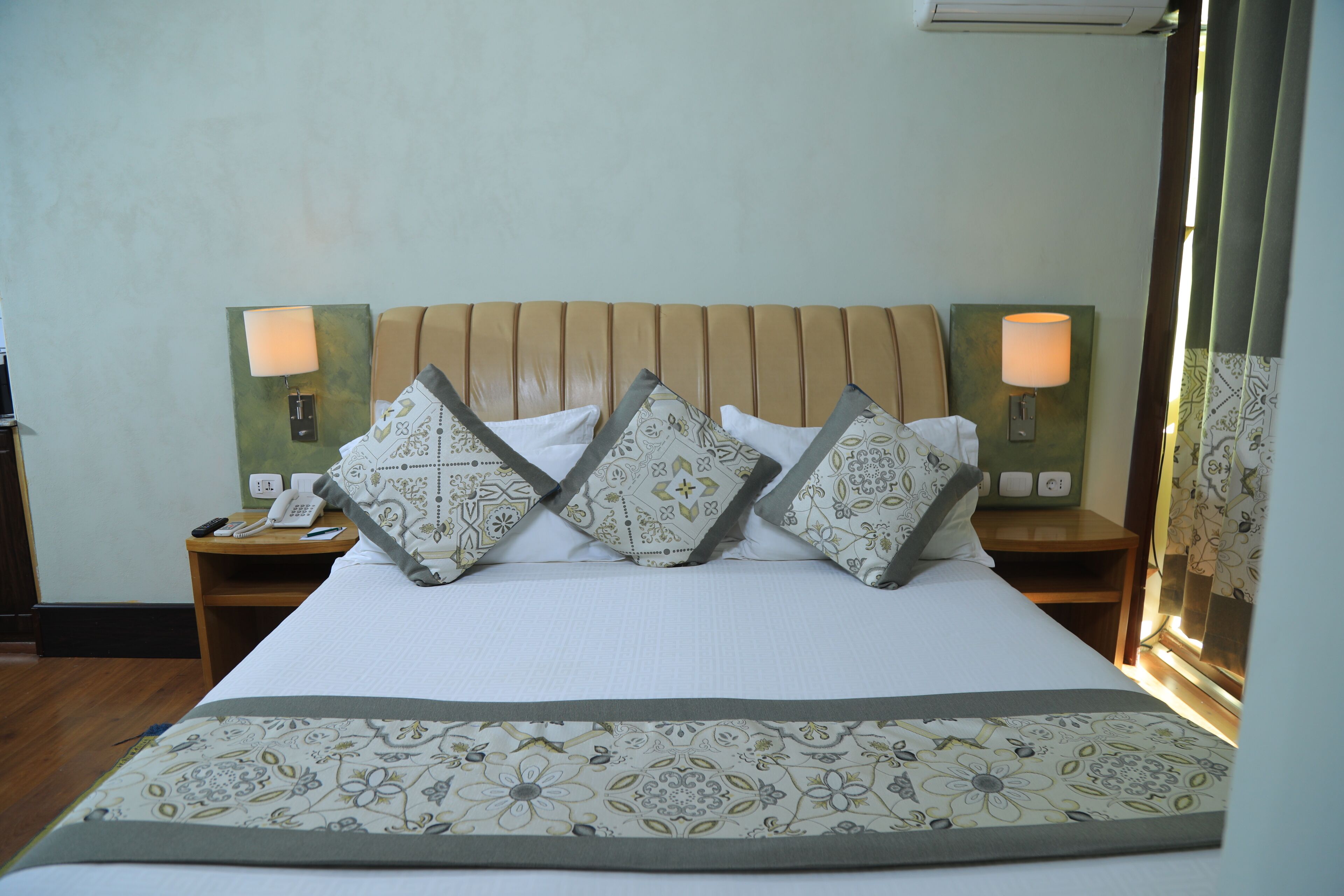 Una cama bien tendida con almohadones estampados y un corredor decorativo. Hay dos mesitas de noche, con lámparas y un teléfono sobre una de ellas.