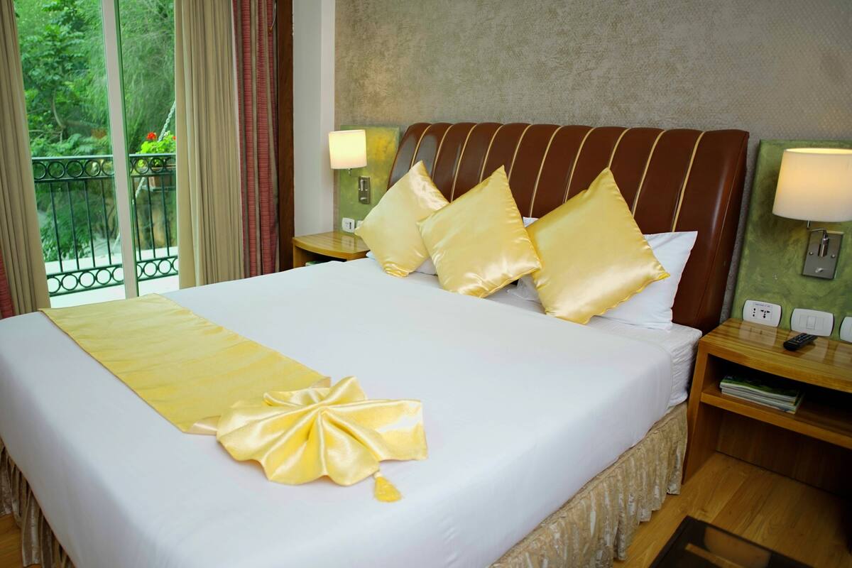 A hotel room with a large bed, two bedside tables, a nightstand, a window with curtains, and a view of greenery outside.