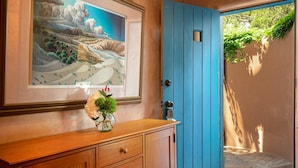 Un mueble de madera con un cuadro enmarcado de un paisaje, un florero con flores y una puerta azul.