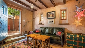 Un cuarto acogedor con un sofá de cuero, una mesa de madera y un tapete colorido. Se ve una ventana con vistas al exterior, una puerta que conduce a otra zona y un baúl verde con detalles metálicos.