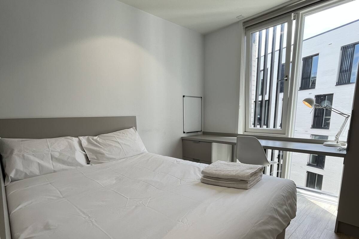 A modern bedroom with a large bed, a desk, and a window with a view of buildings.