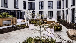 A modern building with a courtyard featuring a concrete pathway, potted plants, and seating areas.