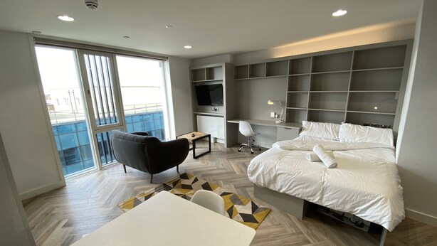 A modern hotel room with a bed, desk, chair, and a dining area.