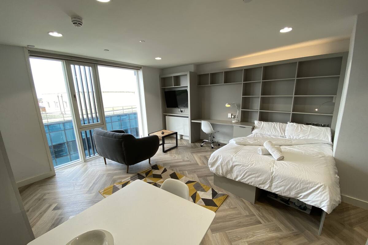 A modern hotel room with a bed, desk, chair, and a dining area.