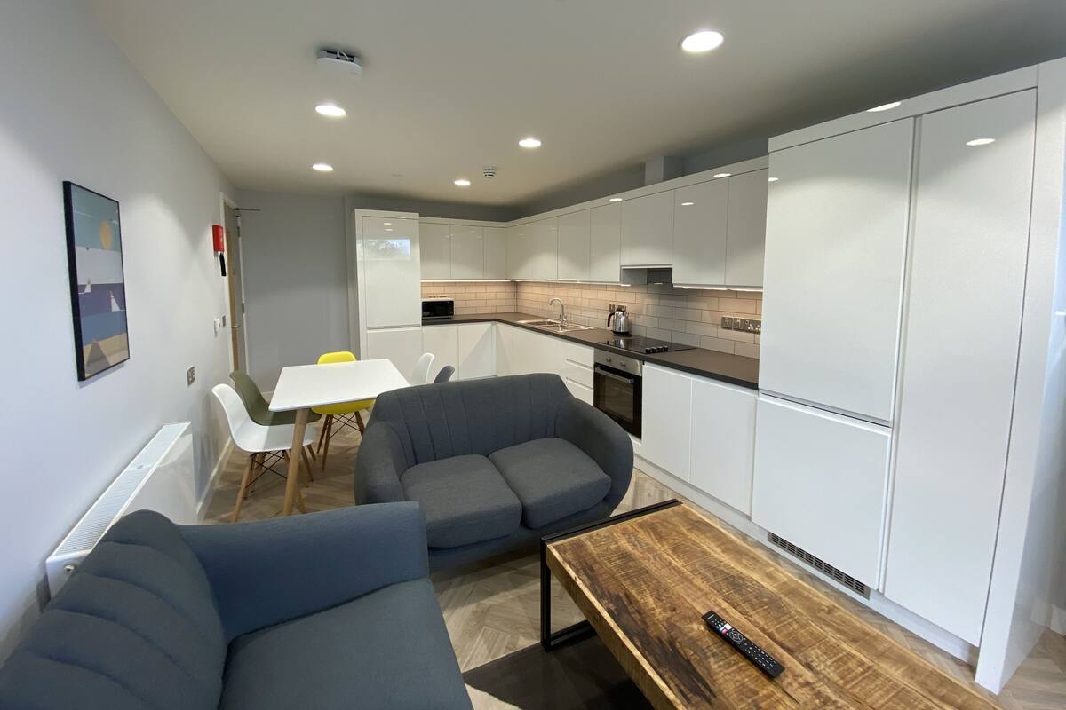 A modern kitchen with white cabinets, a wooden countertop, and a built-in oven. There is a dining area with a white table and two gray sofas.