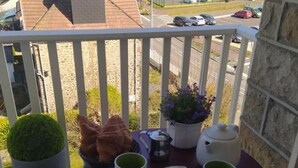 Un balcon avec une table préparée pour le thé, un pot de fleurs et une vue sur une rue bordée de voitures garées.