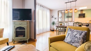 Un salon avec un téléviseur posé sur un meuble, un canapé agrémenté d’un coussin à motifs floraux, une table à manger avec des chaises, et un coin cuisine avec un miroir et des luminaires suspendus.