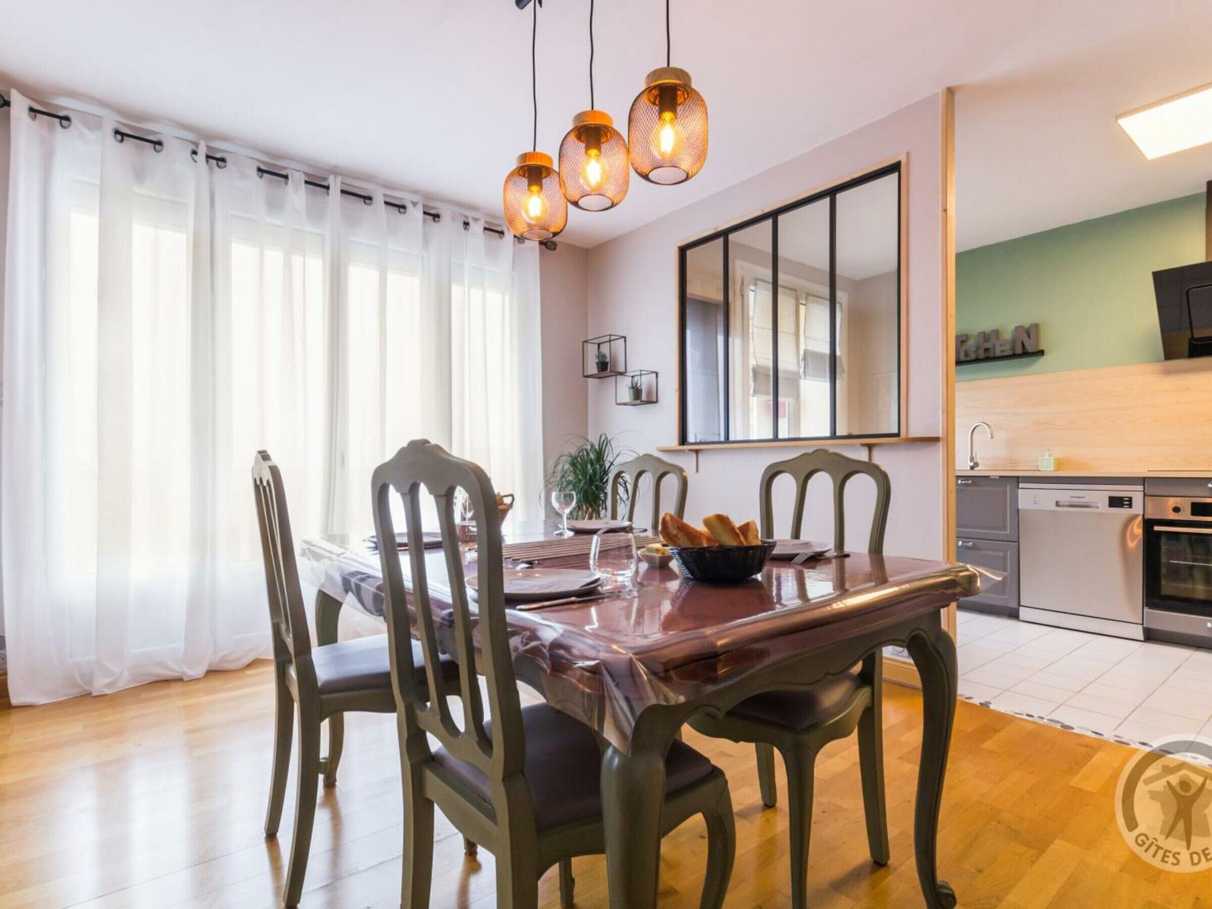 Ein Essbereich mit einem Holztisch, vier Stühlen und einer Kronleuchter. Eine Küchenzeile mit Geschirrspüler und Backofen ist ebenfalls zu sehen.
