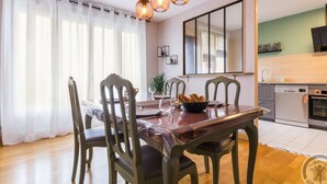 Un coin repas avec une table en bois, quatre chaises et un lustre. On aperçoit également un coin cuisine avec un lave-vaisselle et un four.