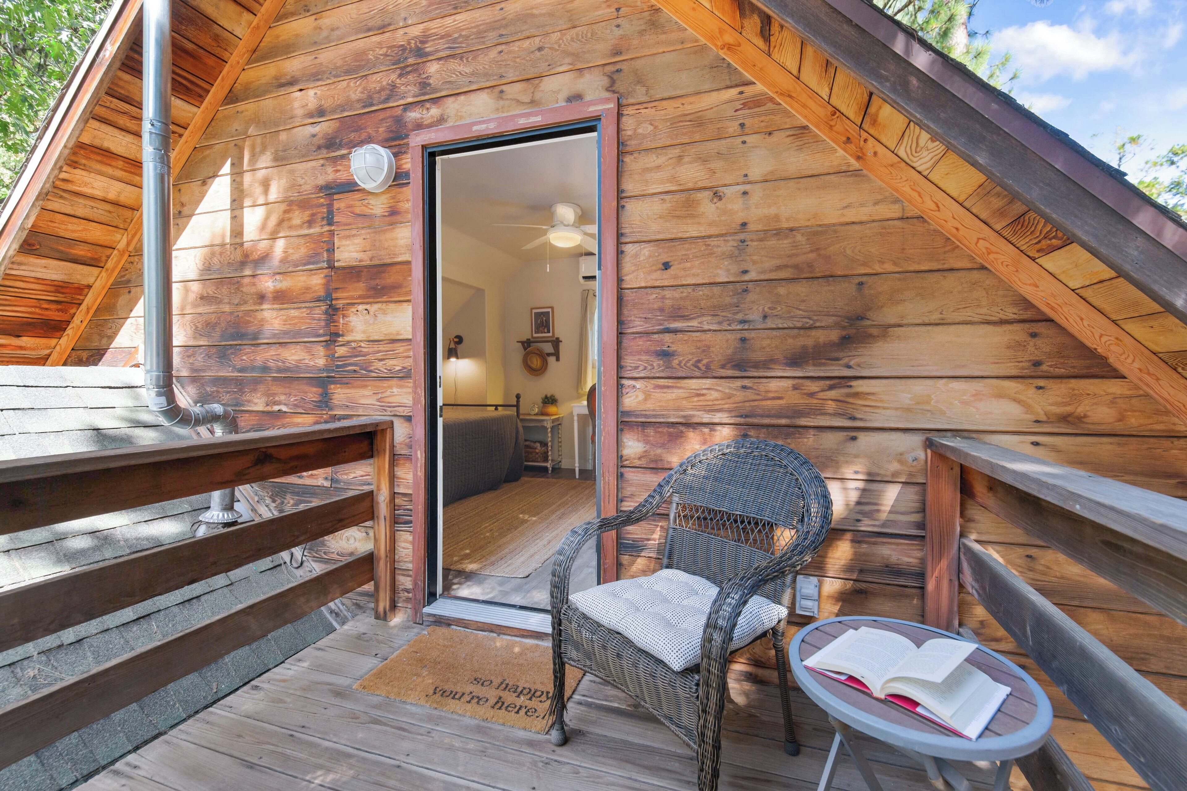 Una baita in legno con un balcone, una sedia in vimini, un tavolino con un libro aperto e una porta d'ingresso.