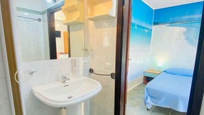 Une salle de bain équipée d’un lavabo, d’un miroir et d’une douche. La pièce adjacente contient un lit recouvert d’une couverture bleue.