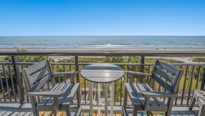 Un balcon avec une table et deux chaises, donnant sur une plage où l’on aperçoit des personnes au loin.
