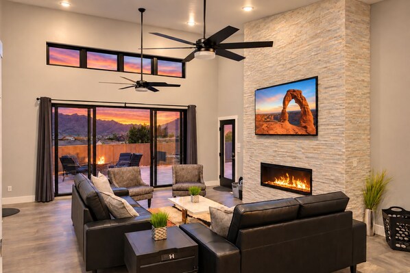 A modern living room with a fireplace, leather sofas, a ceiling fan, and a large window with a scenic view.