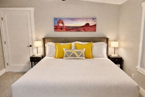 Ein Schlafzimmer mit einem großen Bett, zwei Nachttischen mit Lampen, einer Tür und einem an der Wand hängenden Gemälde einer Naturlandschaft.