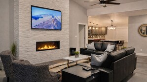 A modern living room with a fireplace, a flat-screen TV mounted on the wall, a ceiling fan, and a dining area in the background. - Moab Desert Escape Villa: Luxury 5-Bedroom Retreat with Heated Pool (Moab)