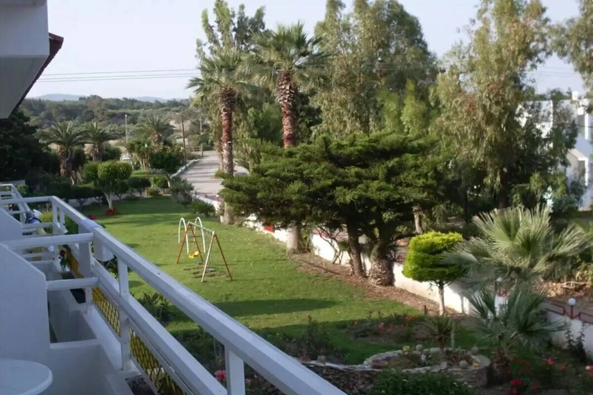 Un balcon doté d’une balustrade blanche, donnant sur un jardin bien entretenu où l’on peut admirer divers arbres et arbustes.