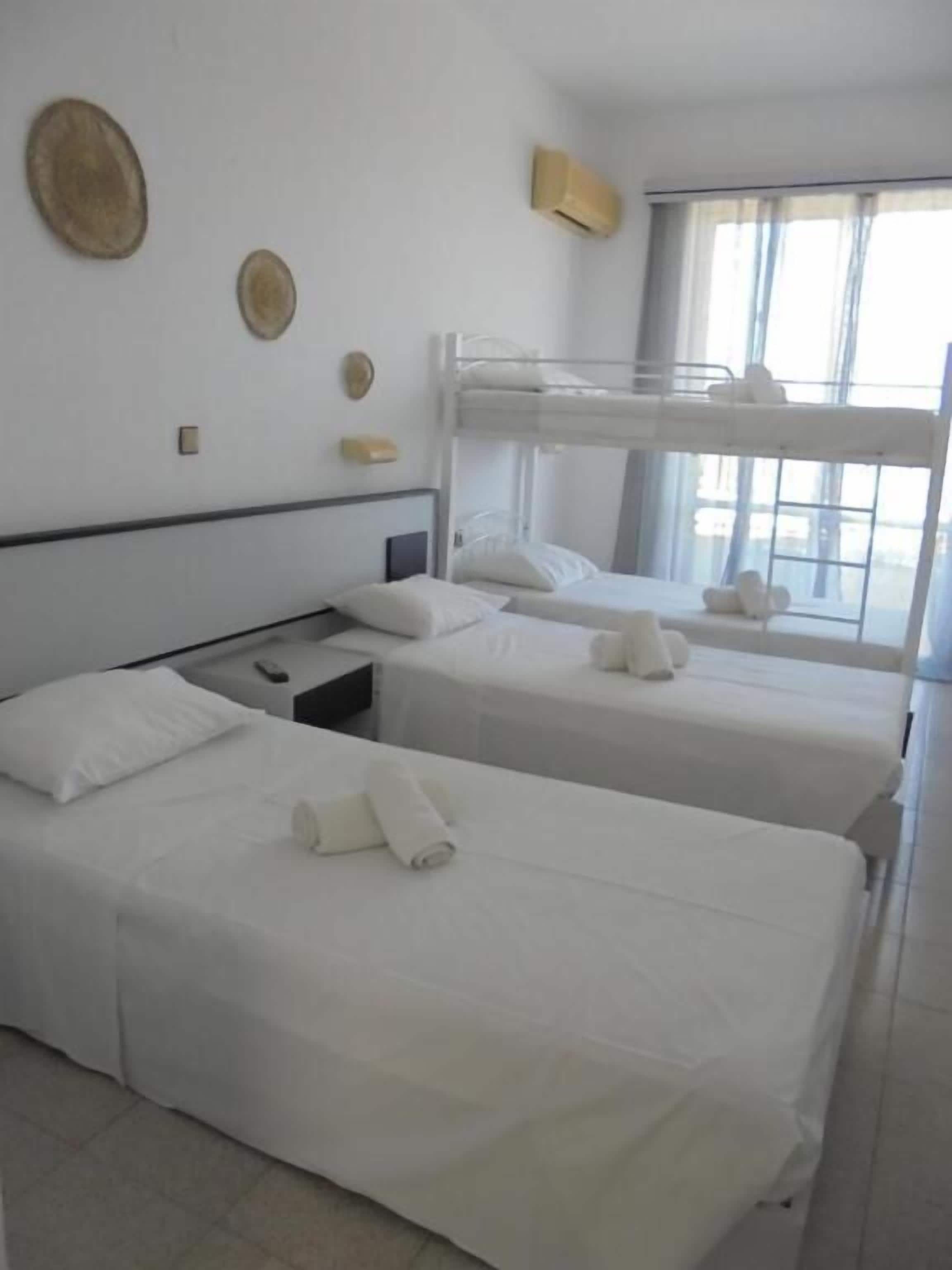 A hotel room with two single beds, a bunk bed, a bedside table with a lamp, a window with curtains, and a wall-mounted air conditioning unit.
