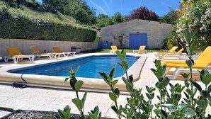 Una piscina rettangolare con bordo in cemento, circondata da poltrone da relax gialle, un siepe e un edificio con una porta blu.