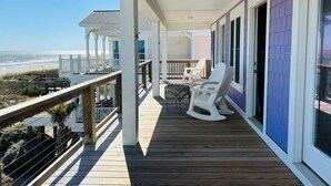 Une terrasse en bois avec une balustrade donnant sur une plage, un espace couvert avec des chaises et une vue sur l’océan.