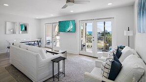A spacious living room with a white sofa, a coffee table, and a TV mounted on the wall. There is a balcony with a view of the ocean and a sailboat. - Private Beach, Pickleball, Tennis 3 heated pools! (Santa Rosa Beach)