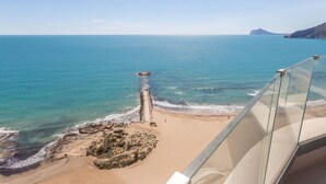 Vista costera desde un balcón con arena dorada, aguas azules cristalinas y un promontorio rocoso.