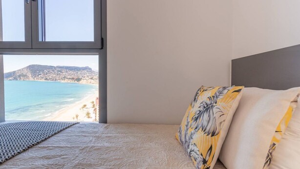 Un dormitorio con cama, ventana con vista a la playa y montañas, y almohada con estampado floral.