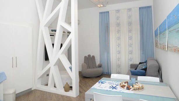 Un salon moderne avec une cloison blanche, un fauteuil gris, un canapé, une table à manger recouverte d’une nappe bleue et un tableau accroché au mur.