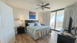A bedroom with a bed, bedside lamps, a ceiling fan, a TV, and a view of the outdoors.