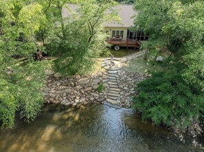 A house with a deck, a stone bridge over a stream, and surrounding greenery.