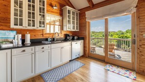 A kitchen with white cabinets, a black countertop, and a wooden ceiling. There is a window with a view of greenery outside. - Shore Enough - Duck, Sanderling, 50 Yds to Beach, Elevator, Pets OK, Community Pool, Flex Stays (Duck)