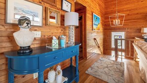 A wooden cabin interior with a blue console table, framed artwork, a chandelier, and a bed. - Shore Enough - Duck, Sanderling, 50 Yds to Beach, Elevator, Pets OK, Community Pool, Flex Stays (Duck)