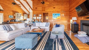 A spacious living room with a large sofa, a striped armchair, and a coffee table. There is a fireplace, a television, and a kitchen area visible in the background. - Shore Enough - Duck, Sanderling, 50 Yds to Beach, Elevator, Pets OK, Community Pool, Flex Stays (Duck)