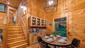 Interior de una cabaña de madera con una mesa de comedor para cuatro personas, una cocina con armarios y una escalera.