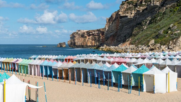 A beach with rows of colorful beach huts and a rocky cliff in the background.