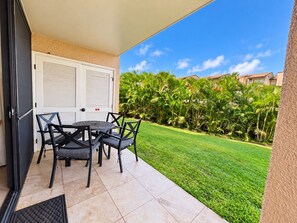 A covered patio with a table and chairs, a door, and a view of a well-maintained lawn and buildings in the background. - Kamaole Sands #9-111 Family-Friendly Suite Near Famous Kamaole III Beach (Kihei)