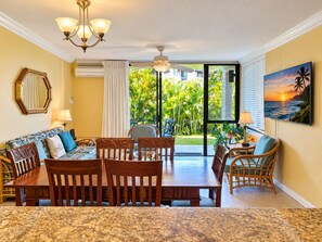 A hotel room with a dining area, a sofa with patterned cushions, a ceiling fan, and a view of a tropical garden.