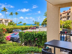 A hotel balcony with a view of the ocean, parked cars, and lush greenery.