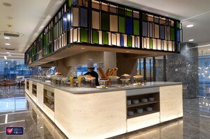 A modern hotel lobby with a central bar area featuring a variety of food and drink options, a seating area with tables, and a large, colorful stained glass window above the bar.