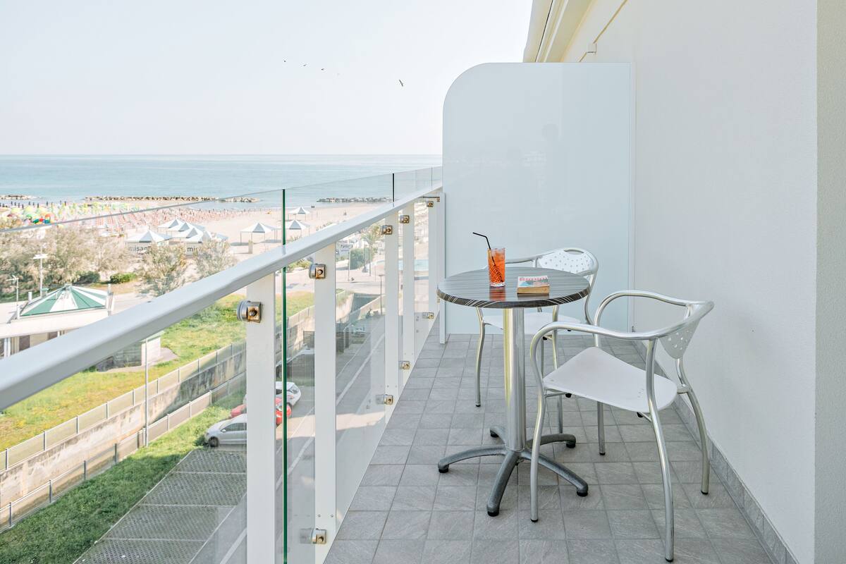 A balcony with a glass railing overlooking a beach, a small table with chairs, and a drink.