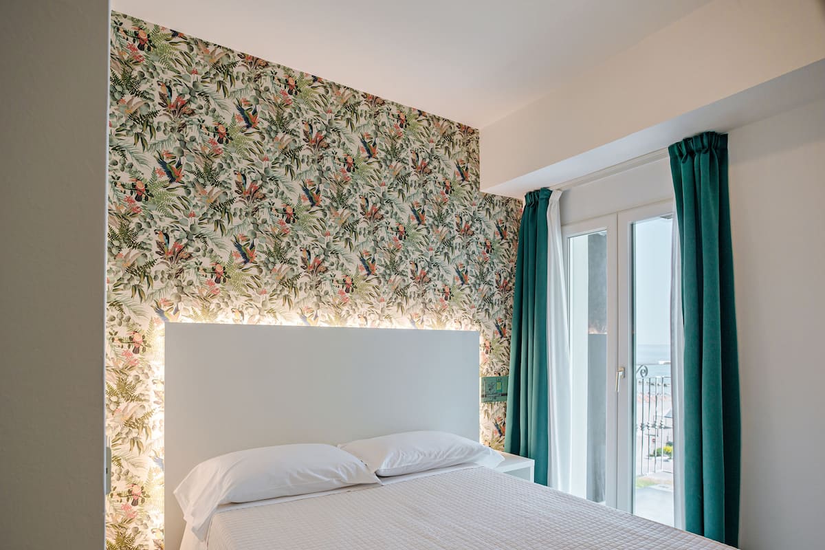 A bedroom with a floral wallpaper, a bed with white bedding, and a view of the outdoors through a window with green curtains.