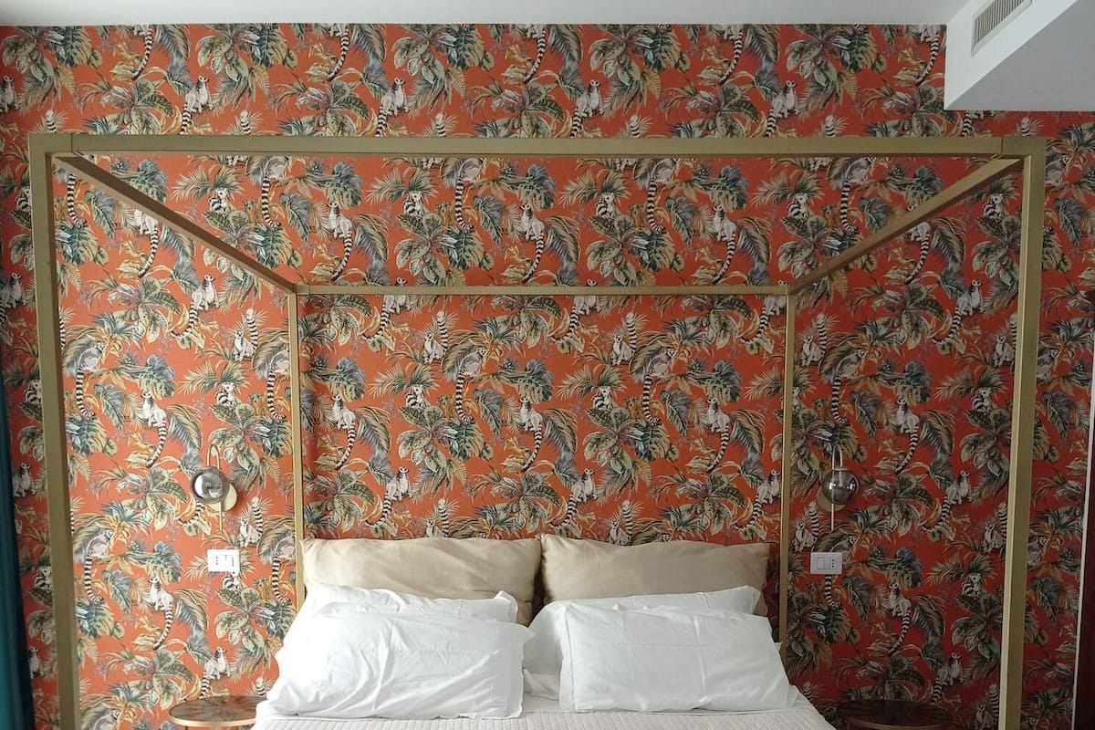 A four-poster bed with white bedding and a wooden frame, set against a wall with tropical wallpaper.