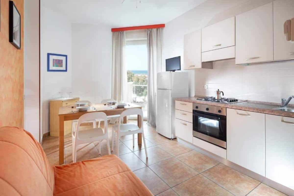 A compact kitchen with white cabinets, a stove, and a refrigerator. There is a dining area with a table and chairs, and a view of the outdoors through a window.