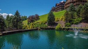 Un lago sereno con una terraza de madera y una fuente, rodeado de abundante vegetación y un albergue con varios balcones.