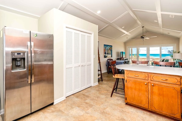 A kitchen with a stainless steel refrigerator, a water dispenser, a wooden cabinet, and a view of the ocean through large windows. - Summer Girl - Oceanfront! 4 Bedroom Cottage (Topsail Beach)